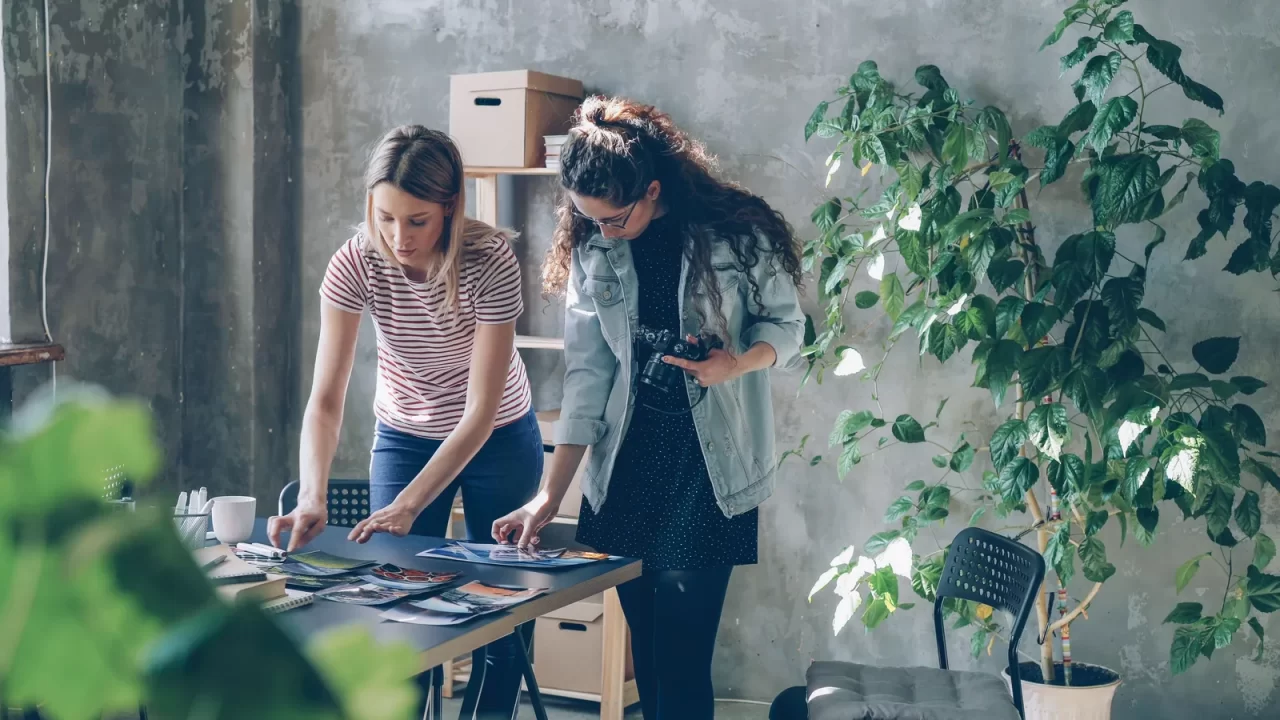 Dos profesionales de una agencia revisan impresiones de trabajos sobre una mesa para elegir qué proyectos mostrar a clientes calificados