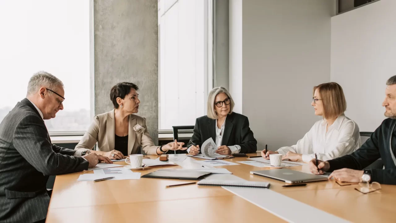Executivos reunidos em sala de reunião discutem papéis sobre a mesa, avaliando o custo e o retorno de buscar reuniões com grandes contas